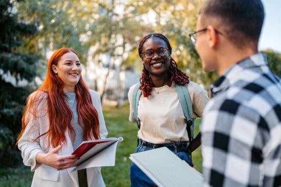 Three young adults standing outside, smiling and talking, holding notebooks and folders.