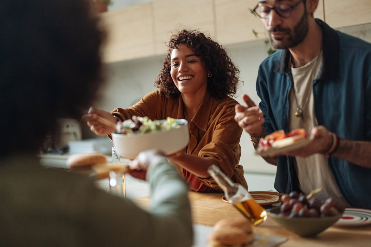 Smiling woman passes a bowl of salad at a kitchen table with friends enjoying food and drinks.