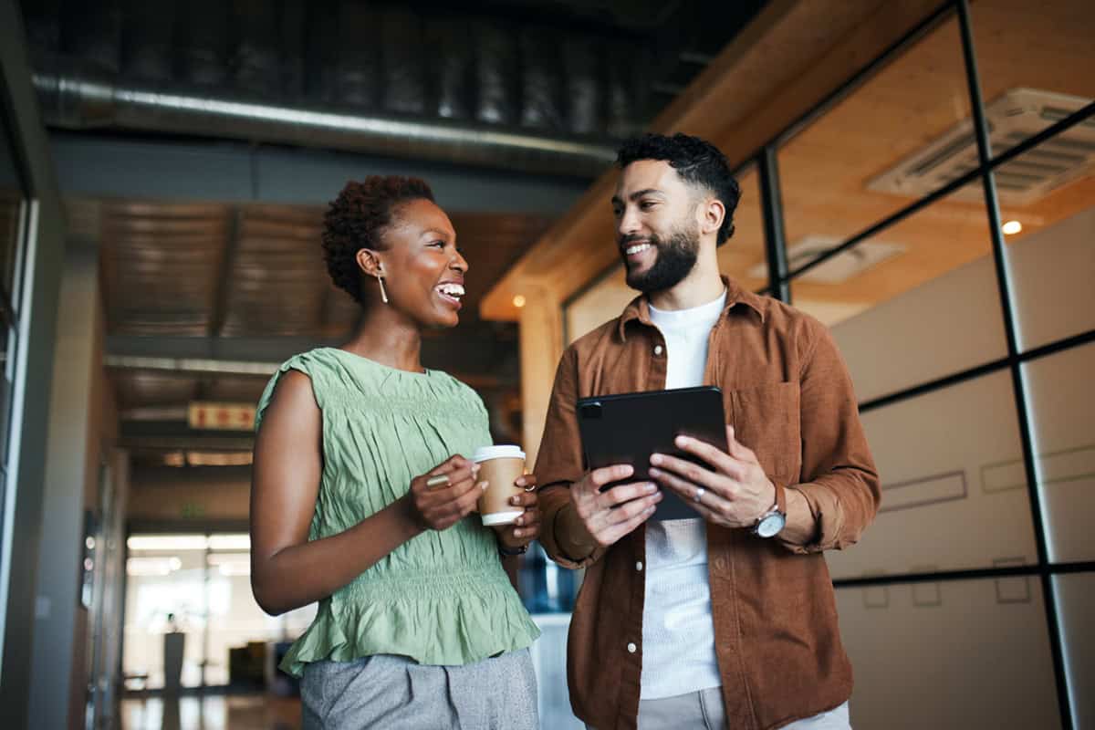 Two coworkers smiling and talking in an office; one holds a coffee cup, the other a tablet.