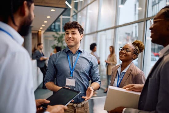 Three professionals with badges smile and talk in a modern office hallway, holding folders and a tablet.