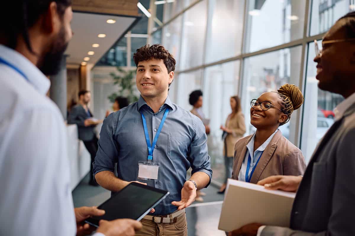 Three professionals with badges smile and talk in a modern office hallway, holding folders and a tablet.