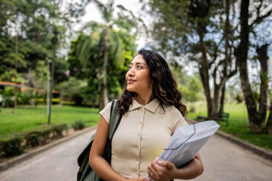 Woman with wavy hair holding books walks outdoors in a park, looking to the side and smiling.