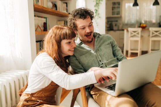 A smiling couple sits together, looking excitedly at a laptop screen in a bright, cozy room.