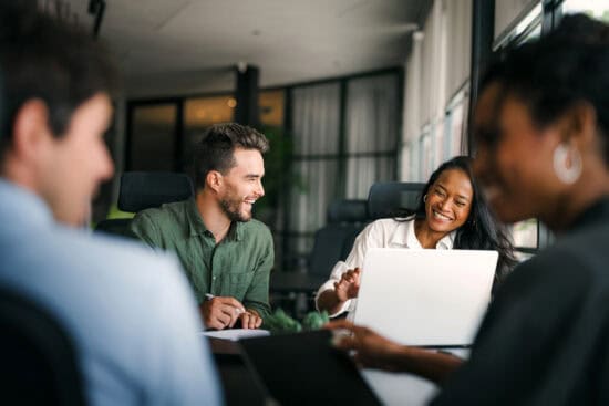 Four people sit around a table in an office, smiling and talking, with laptops and notebooks in front of them.