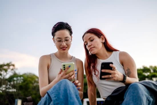 Two women sitting outdoors, looking at their phones and smiling, with trees and sky in the background.