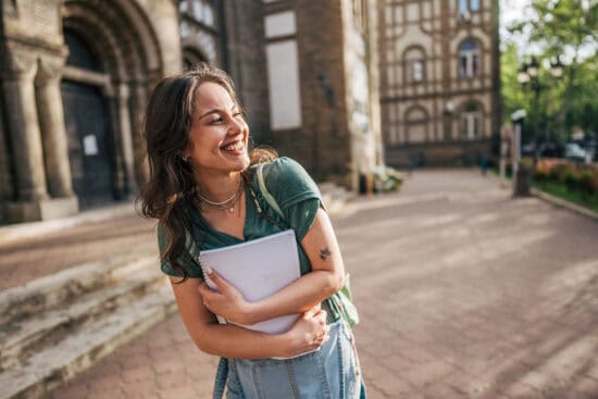 Smiling young woman holding a laptop stands outside a historic building on a sunny day.