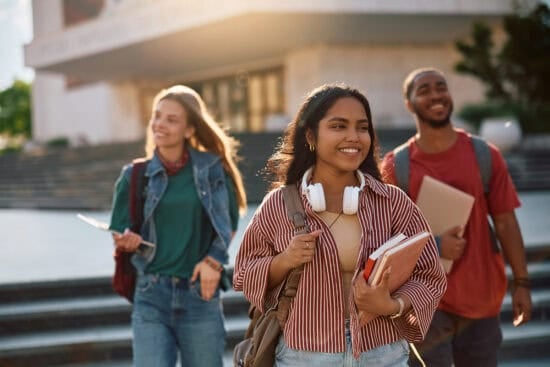 Three smiling students with books and backpacks walk outside on a sunny day near a building with steps.