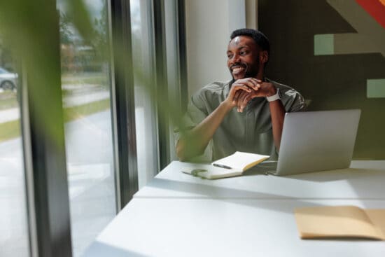 Smiling man sitting at a desk with a laptop, notebook, and pen, looking out a window.