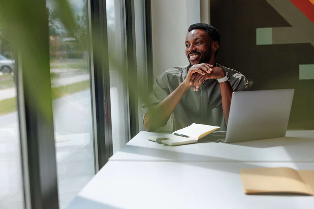 Smiling man sitting at a desk with a laptop, notebook, and pen, looking out a window.