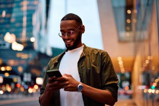 A man smiling while using his phone on a city street at dusk, with blurred lights in the background.