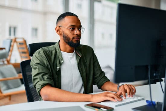 Man with glasses and beard working on a computer at a modern office desk with a phone beside him.