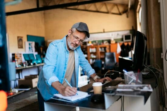 Older man in a cap writes in a notebook while working at a desk with a computer in a workshop.