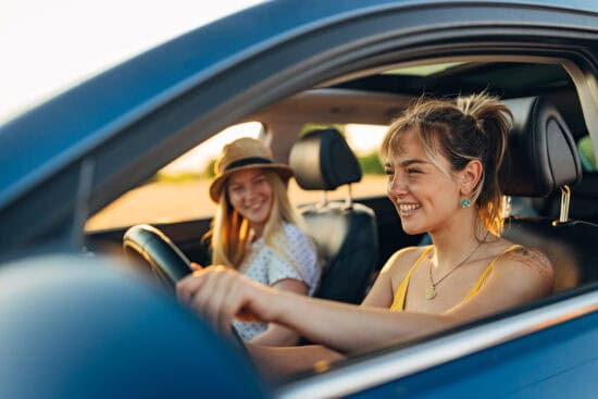 Two young women smiling in a car, one driving and the other in the passenger seat on a sunny day.