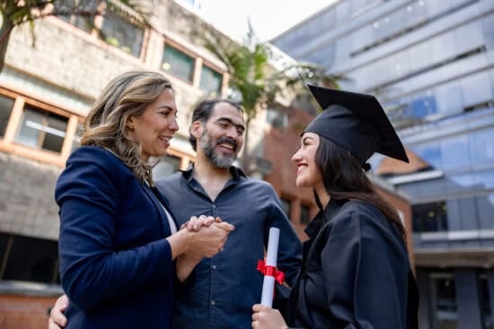 A graduate in cap and gown smiles with two adults, holding a diploma outdoors near modern buildings.