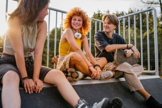 Three smiling teens sitting together at a skatepark, chatting and relaxing next to a metal railing.