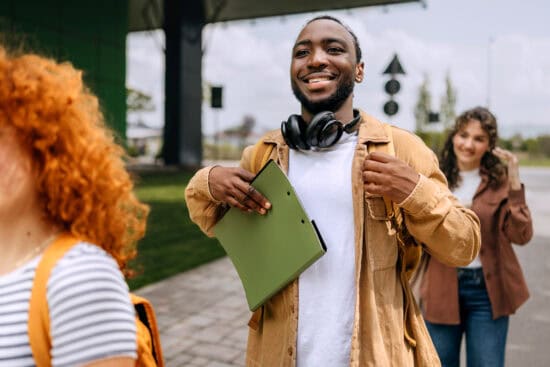 Smiling student holding folders and wearing headphones walks outdoors with friends.