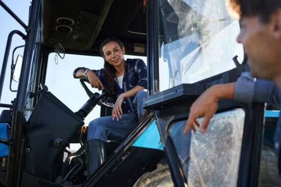 A young woman sits in a blue tractor cab, smiling at a man standing outside the vehicle.