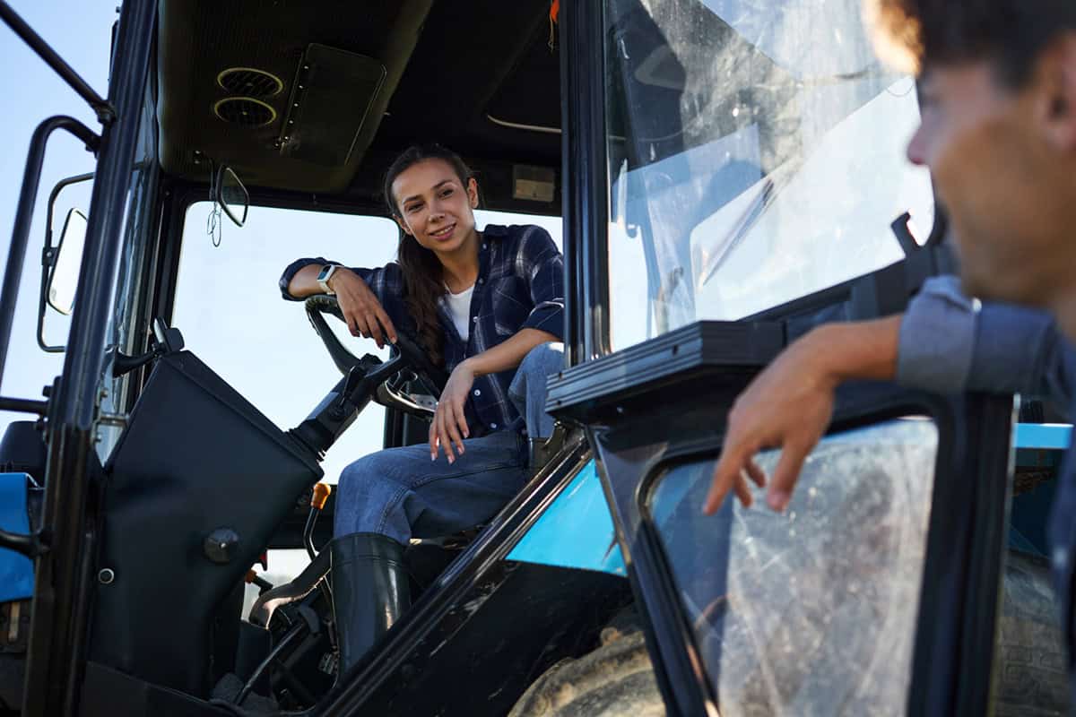 A young woman sits in a blue tractor cab, smiling at a man standing outside the vehicle.