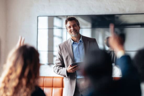 Smiling man in a suit holding a tablet, engaging with people raising hands in an office setting.