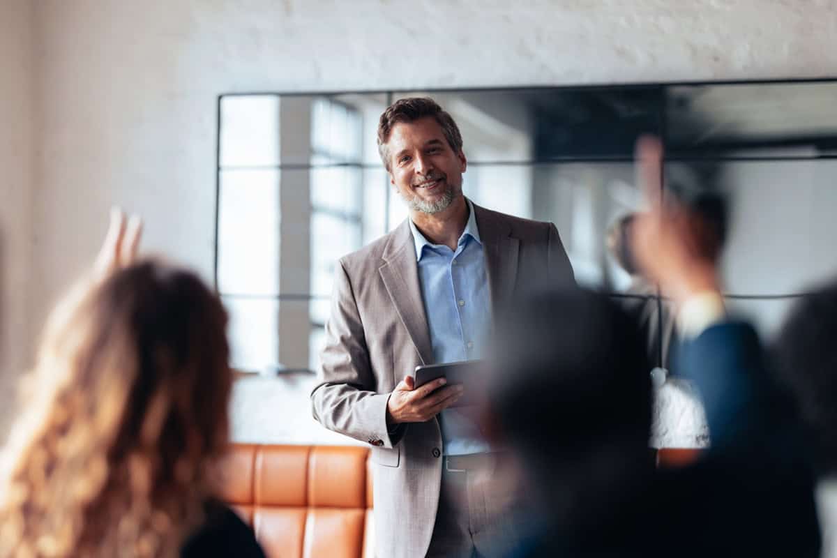 Smiling man in a suit holding a tablet, engaging with people raising hands in an office setting.