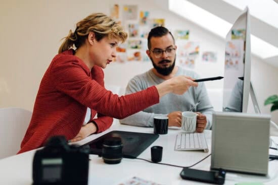 Woman pointing at computer screen while man watches; both seated at desk with coffee cups and camera equipment.