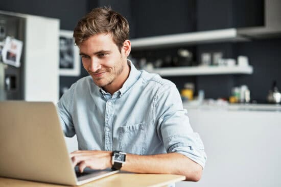 Man in a light blue shirt smiling while using a laptop at a kitchen table.