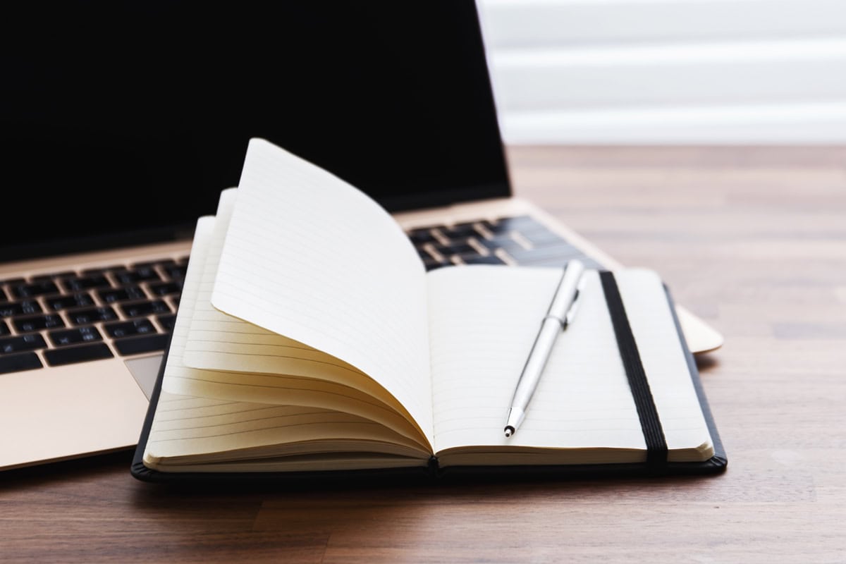 Open notebook with a pen resting on it, placed on a laptop keyboard on a wooden desk.
