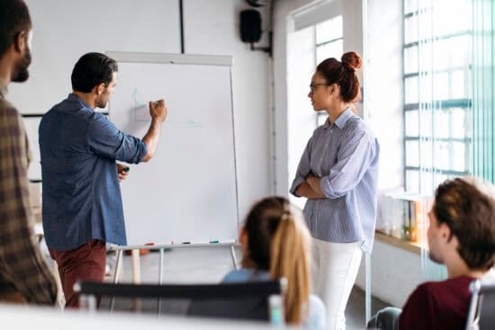 A man draws on a whiteboard while colleagues watch and listen during a meeting in a bright office.