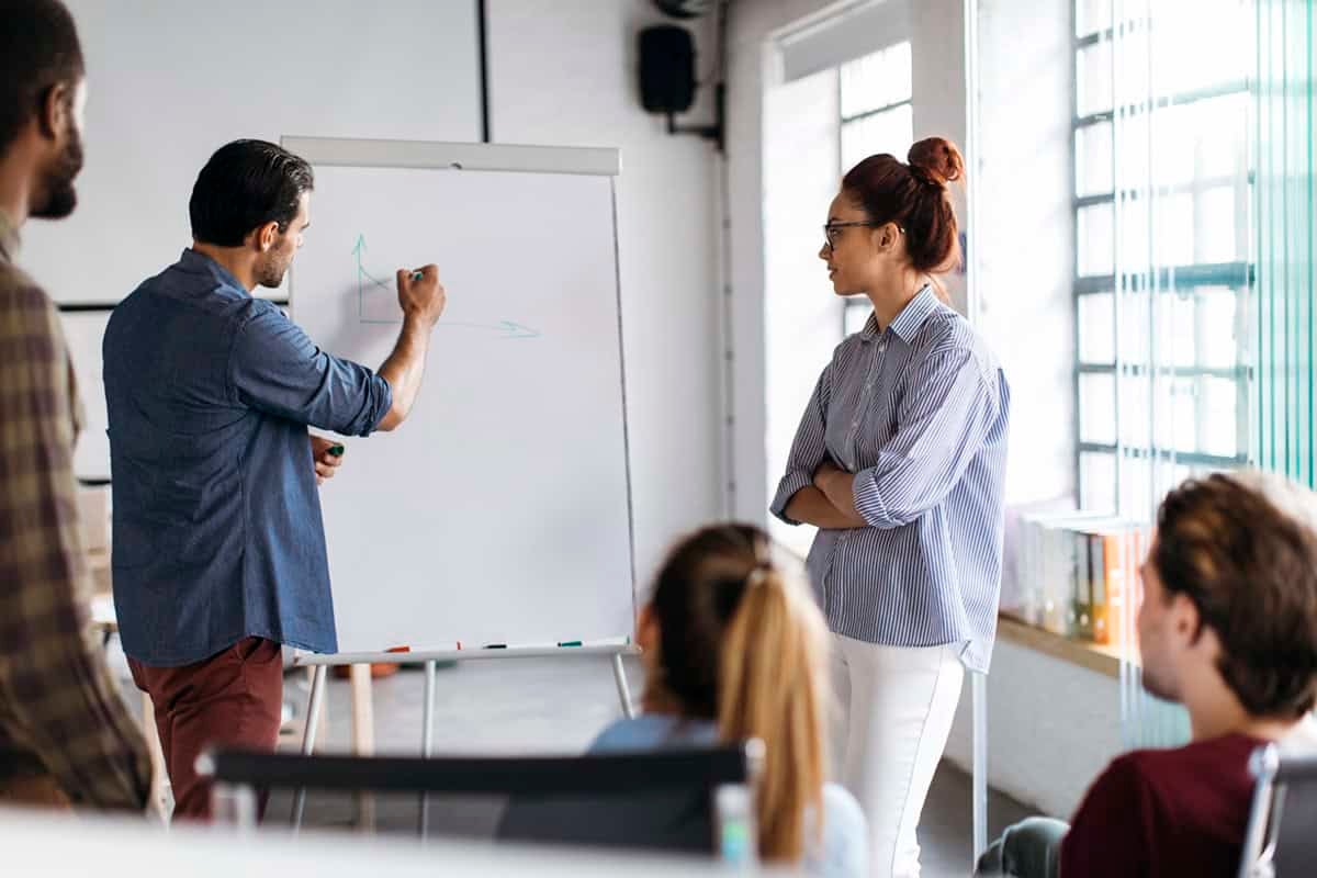 A man draws on a whiteboard while colleagues watch and listen during a meeting in a bright office.