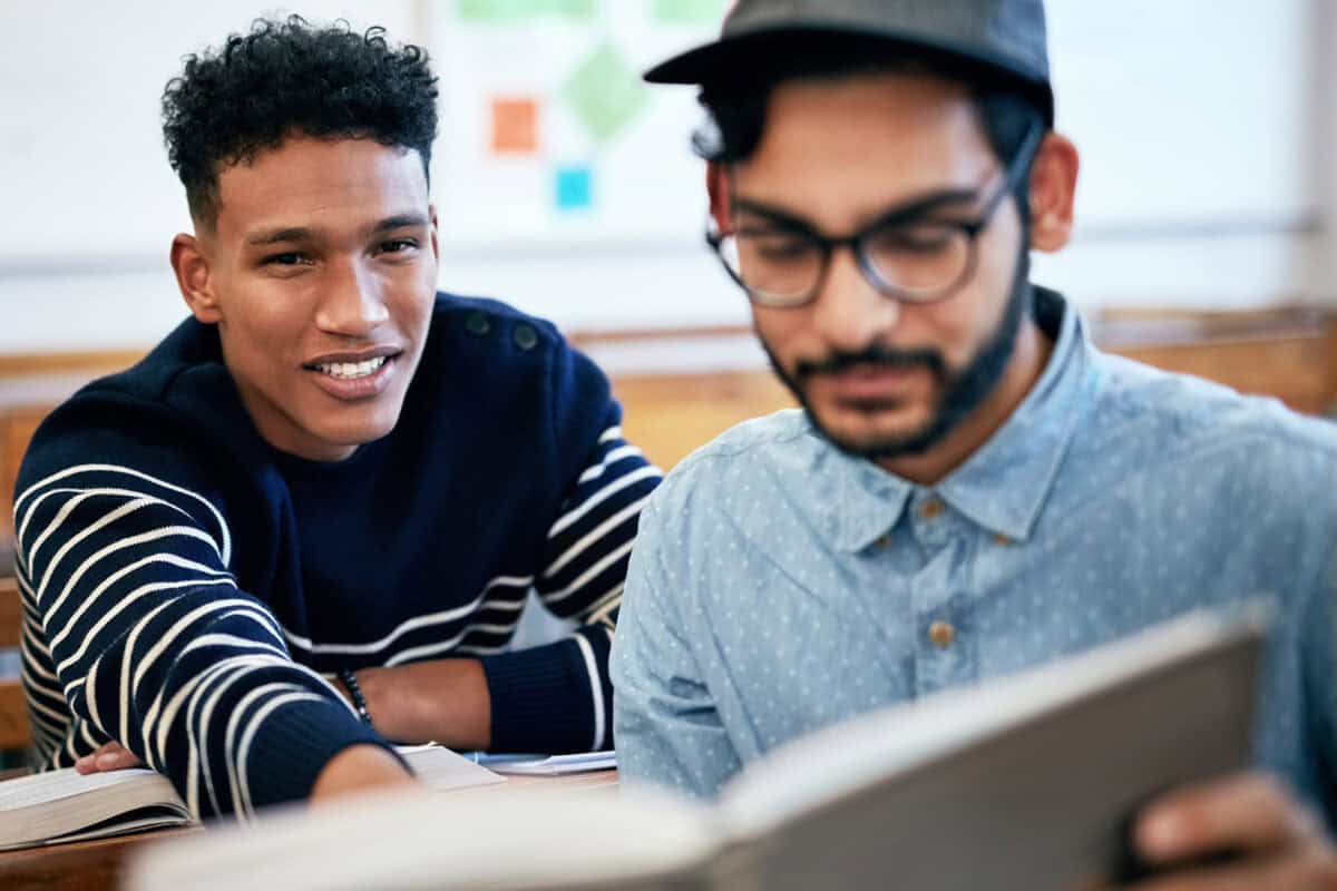 Two young men sitting together in a classroom, one reading a book while the other smiles at the camera.
