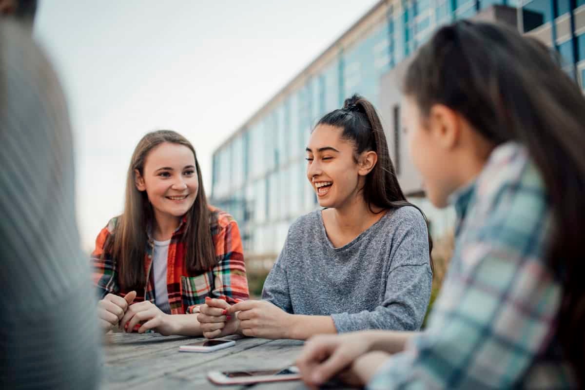 Three teenage girls sit at a table outside, smiling and talking, with a modern building in the background.