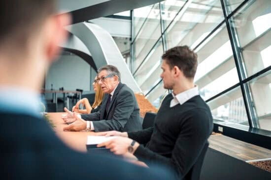People in business attire sit at a table in a modern office with large windows, engaged in discussion.
