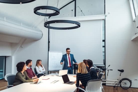 A man in a suit presents to colleagues around a conference table in a modern office meeting room.