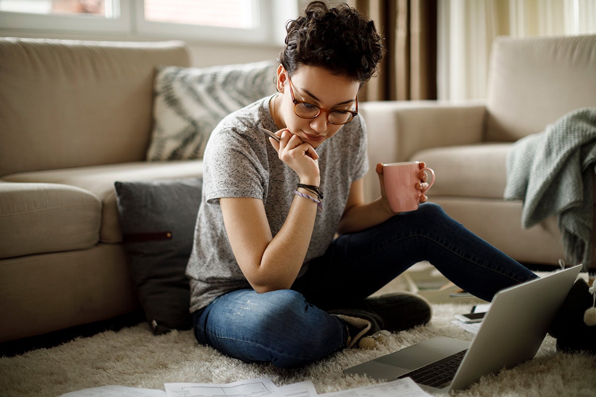 Person sitting on floor, holding a mug, looking thoughtfully at papers and a laptop in a cozy living room.