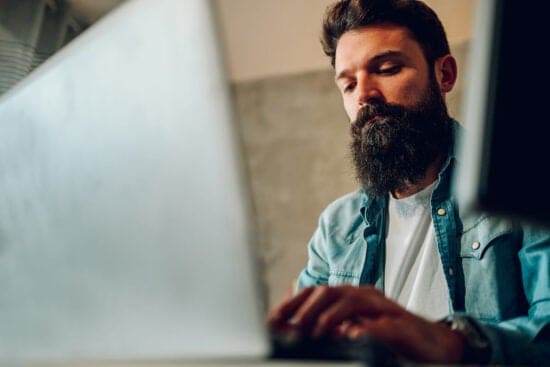 Man with a beard and denim shirt working on a laptop at a desk, focused on the screen.