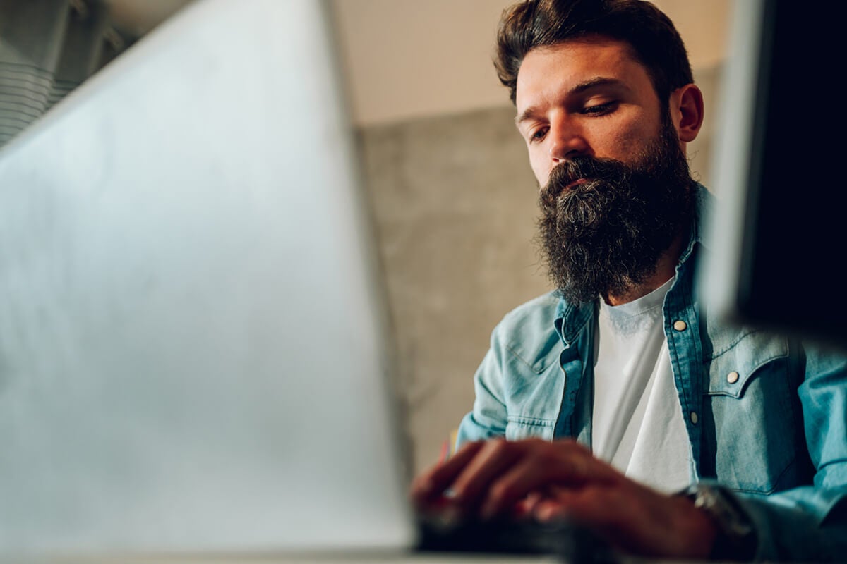 Man with a beard and denim shirt working on a laptop at a desk, focused on the screen.