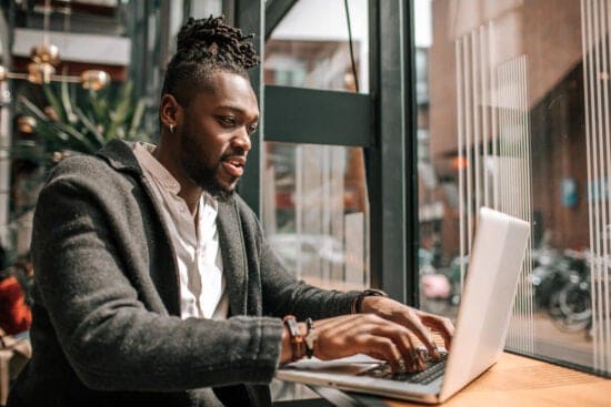 Man in a coat working on a laptop at a table near large windows in a modern cafe or office.
