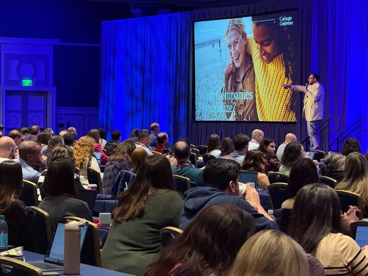 A speaker presents to a seated audience in a conference room, with a large screen displaying a slide.