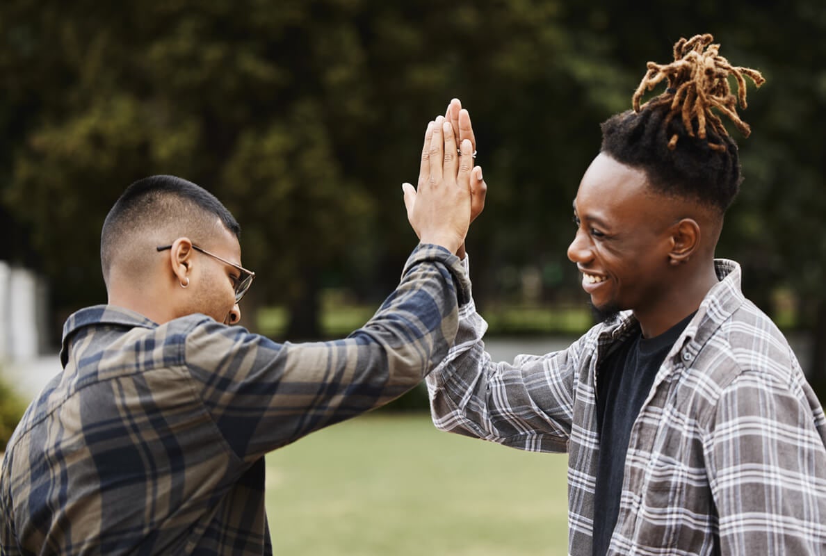 Two young men outdoors giving each other a high five and smiling, both wearing plaid shirts.