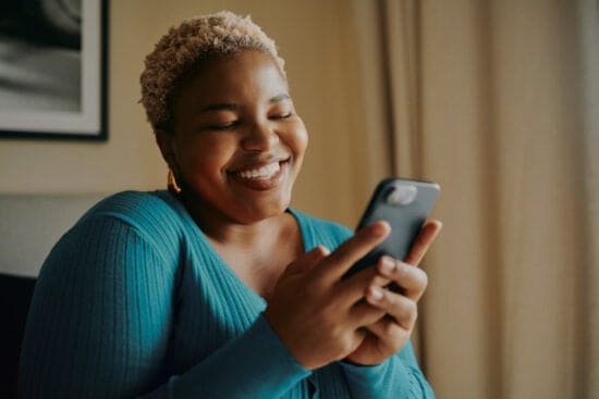 Smiling woman in a teal sweater holds and looks at her smartphone indoors.
