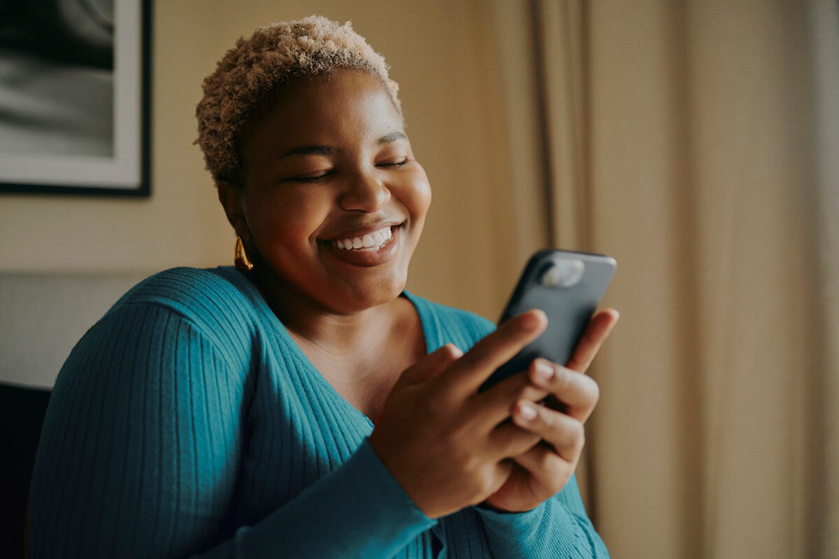 Smiling woman in a teal sweater holds and looks at her smartphone indoors.