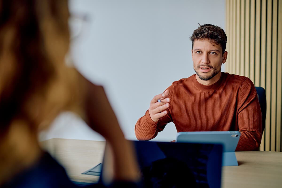 Man in brown sweater talking to a woman across a desk in an office setting, with a tablet in front of him.