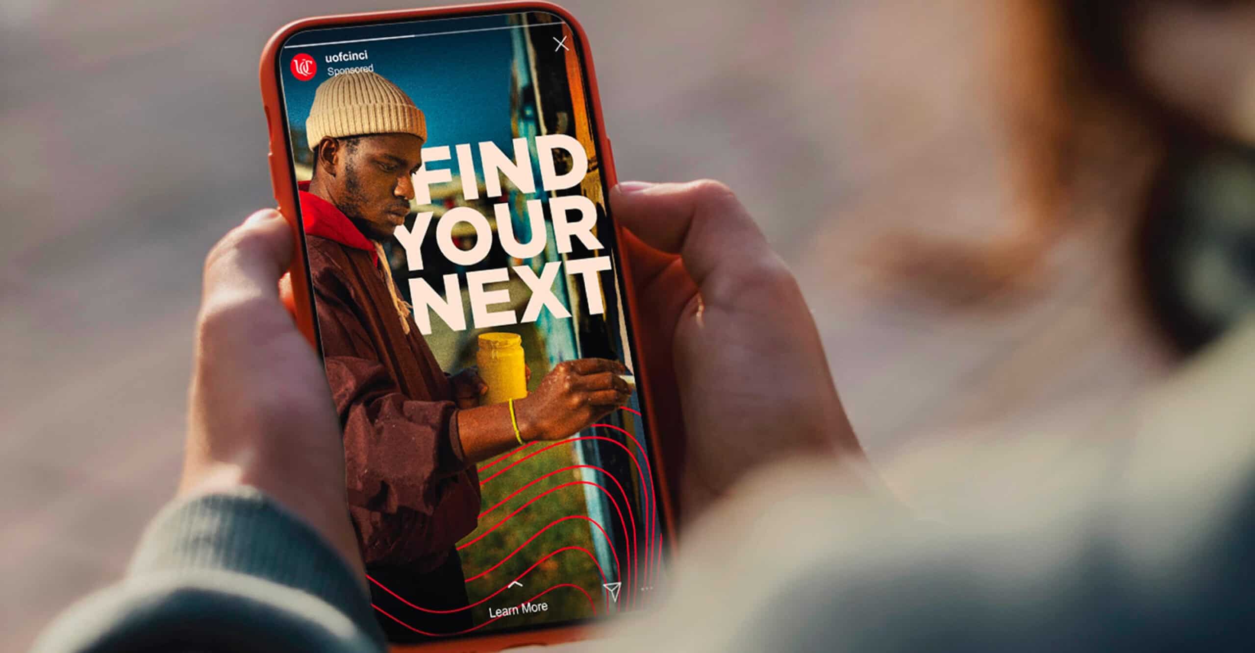 Person holding a phone displaying an ad with "FIND YOUR NEXT" over an image of a man in a beanie.