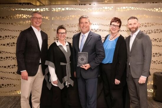 Five people in business attire stand smiling; the man in the center holds a plaque or award.