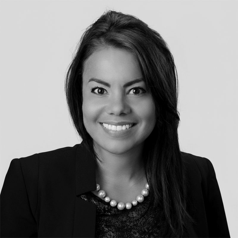 Smiling woman with long hair, wearing a blazer, pearl necklace, and dark top, on a plain background.