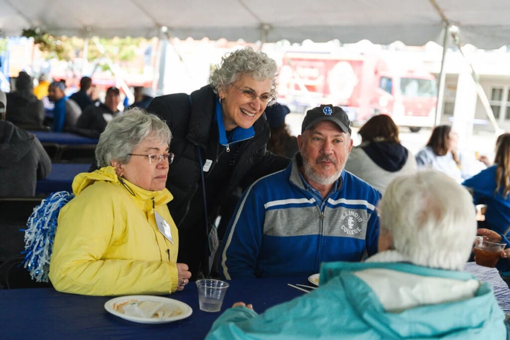 Four older adults sit and talk at a table under a tent during an outdoor event, with food on the table.