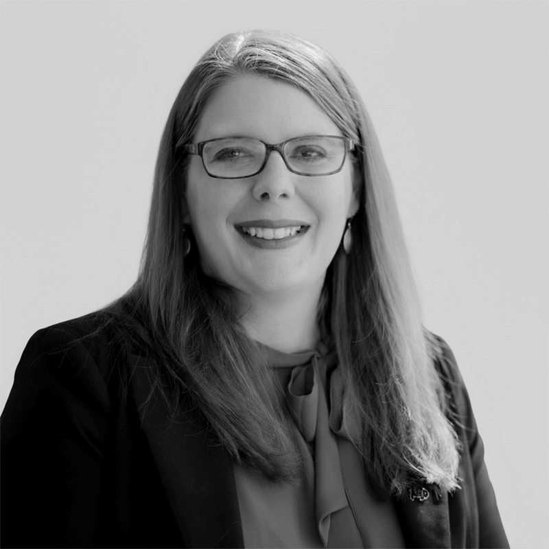 A woman with long hair and glasses, smiling, wearing a blazer and blouse, in a black and white photo.
