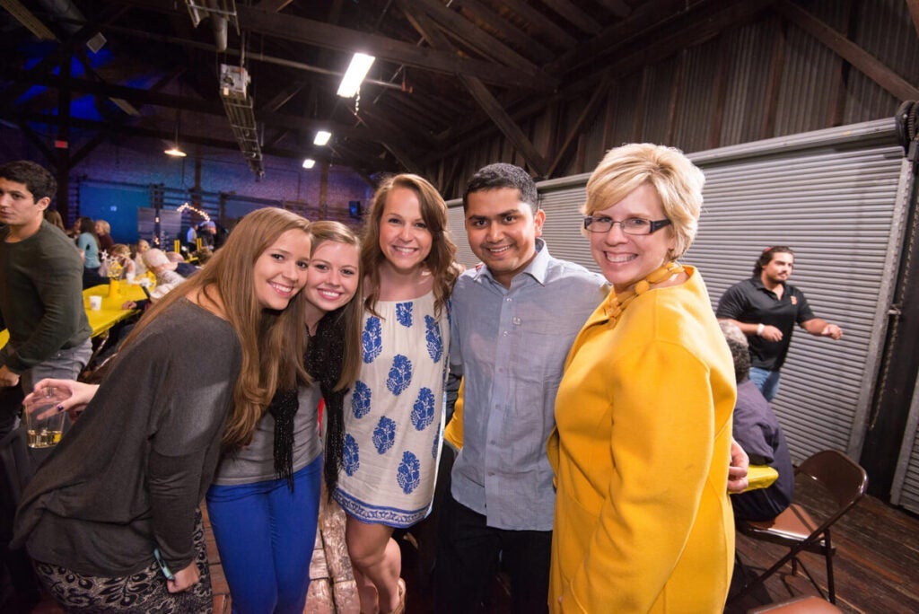 Five people smiling and posing together at an indoor event with tables and others in the background.