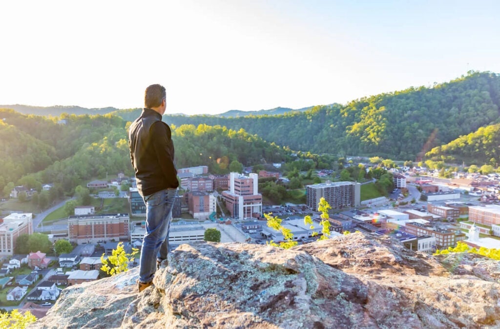Man standing on a rocky cliff overlooking a town surrounded by green hills in bright sunlight.
