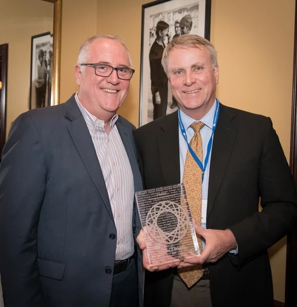 Two men in suits smiling; one is holding a clear engraved award or plaque.